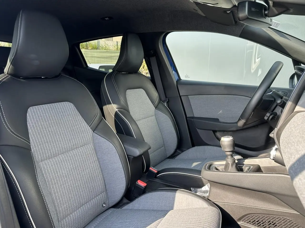 Intérieur avant de la Renault Clio TCE 90 Techno 2025, sièges tissu gris et noir, volant multifonction et levier de vitesse manuel.