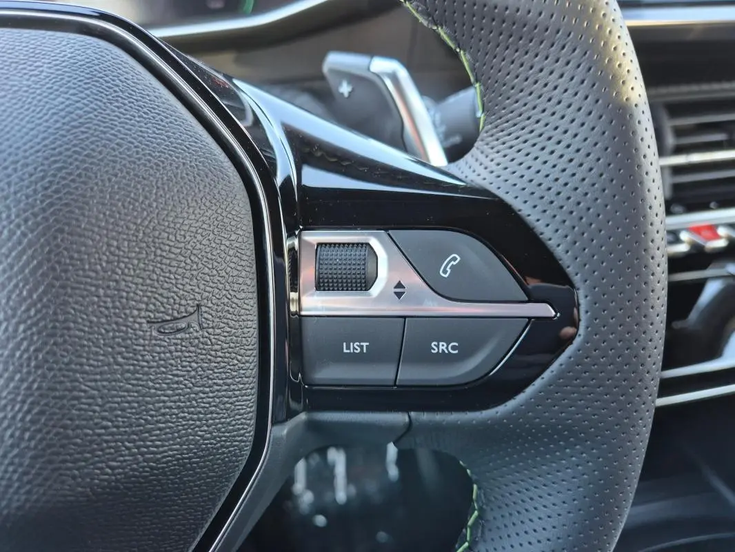 Gros plan sur le côté gauche du volant perforé noir de la Peugeot 208 Hybrid 136 e-DCS6 GT avec commandes audio intégrées.