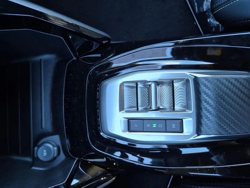 Gros plan sur la console centrale noire brillante de la Peugeot 208 Hybrid 136 e-DCS6 GT avec commandes de boîte auto et prise 12V.