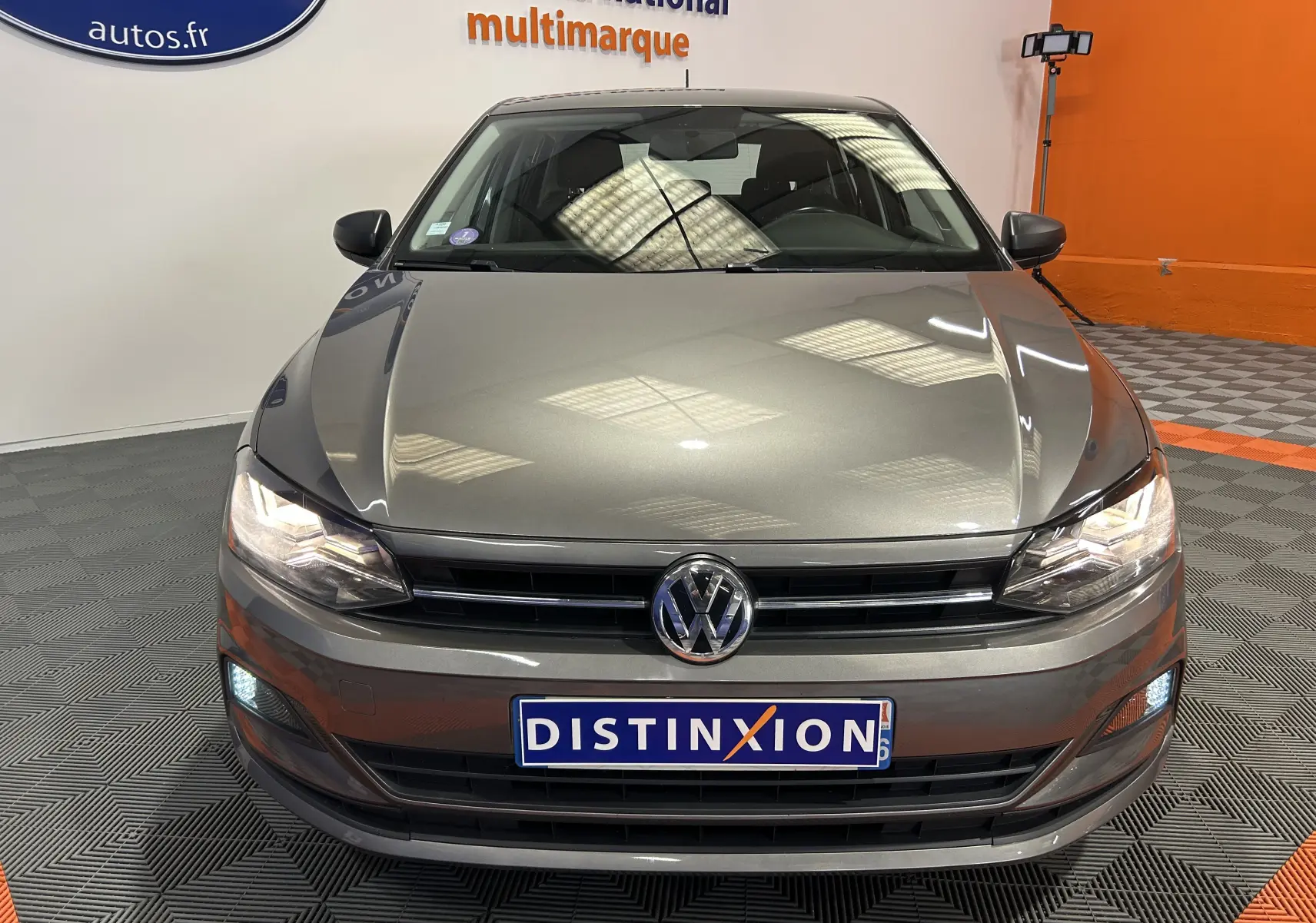 Vue avant d'une Volkswagen Polo gris métallisé avec phares allumés dans un showroom au sol gris et orange.