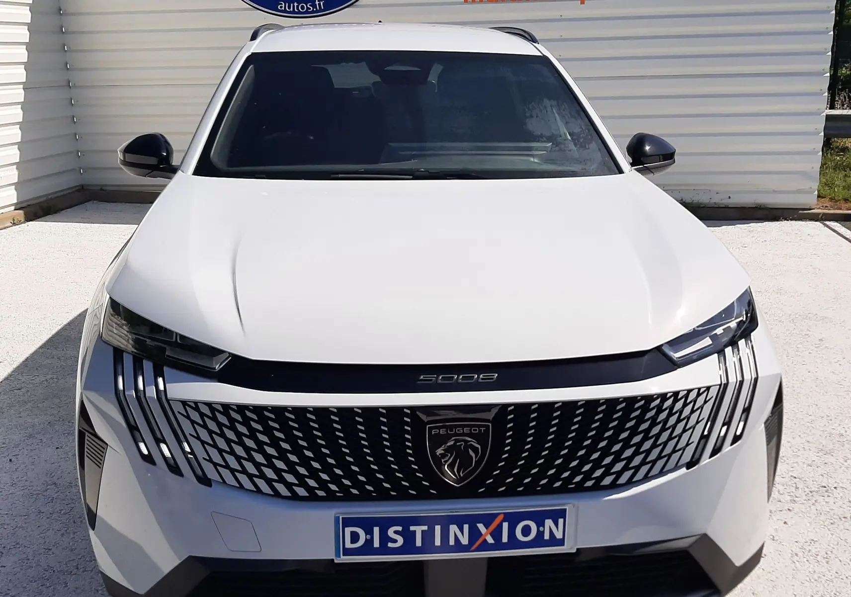Vue avant d'un Peugeot 5008 blanc Okenite avec calandre noire distinctive et logo lion central, stationné devant un mur publicitaire.