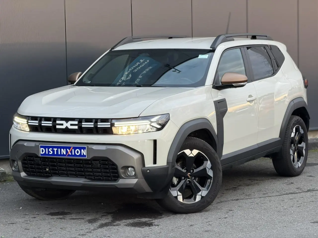 Dacia Duster beige Sandstone en 3/4 avant droit avec jantes alliage noires et barres de toit grises.