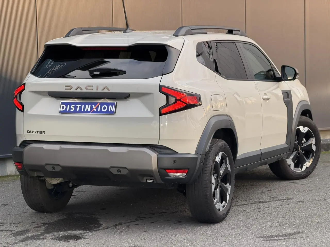 Vue 3/4 arrière droite du Dacia Duster beige Sandstone avec jantes alliage et barres de toit noires.