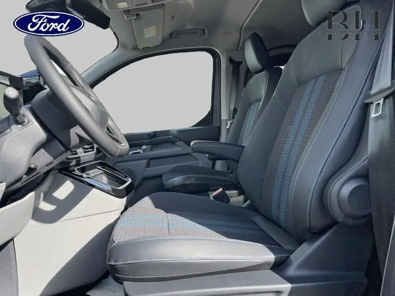 Vue intérieure côté conducteur du Ford Transit Custom Fourgon 2026, sièges tissu noir avec surpiqûres bleues et accoudoir.