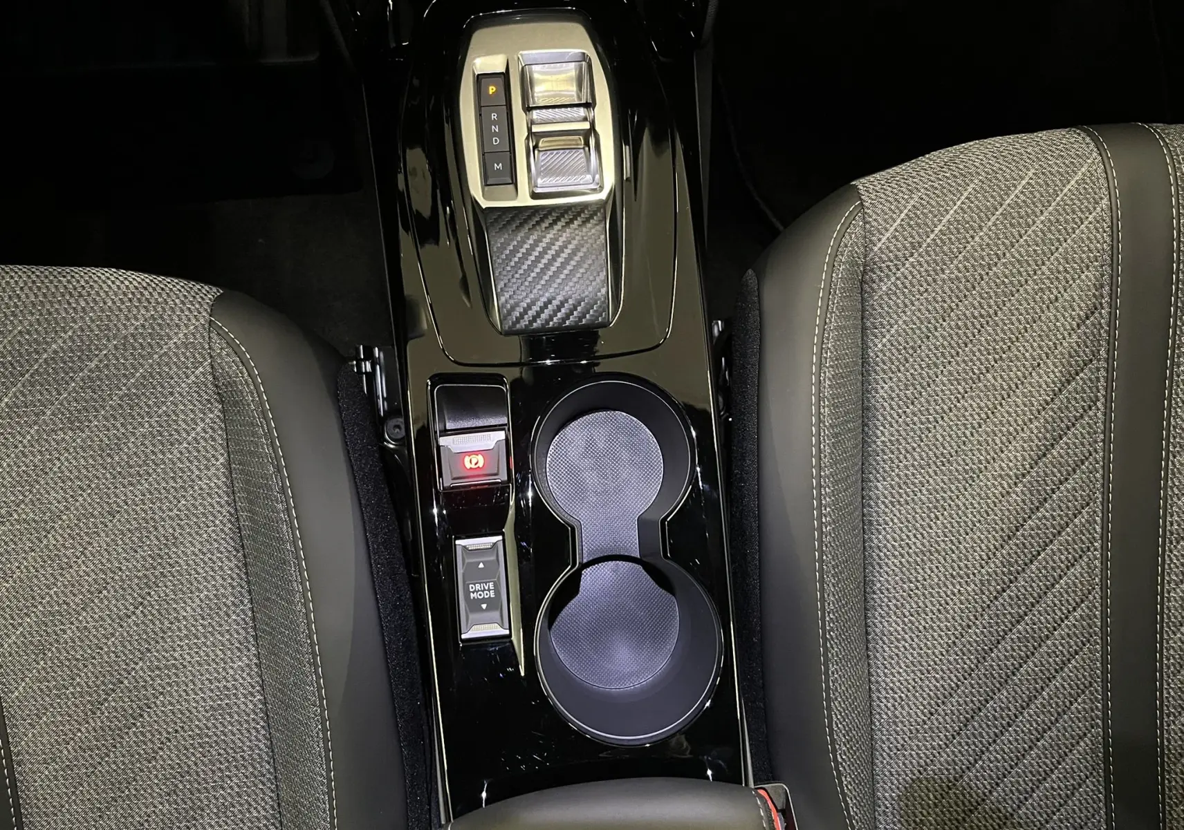 Vue plongeante sur la console centrale noire brillante du Peugeot 2008 2025, avec sélecteur de vitesse et porte-gobelets entre sièges gris tissu.
