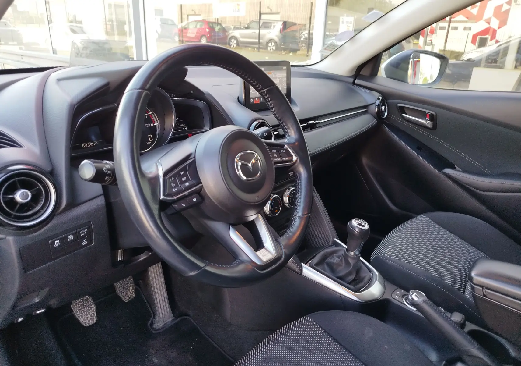 Vue intérieure côté conducteur de la Mazda2 2018, volant cuir, levier de vitesse manuel et tableau de bord moderne.