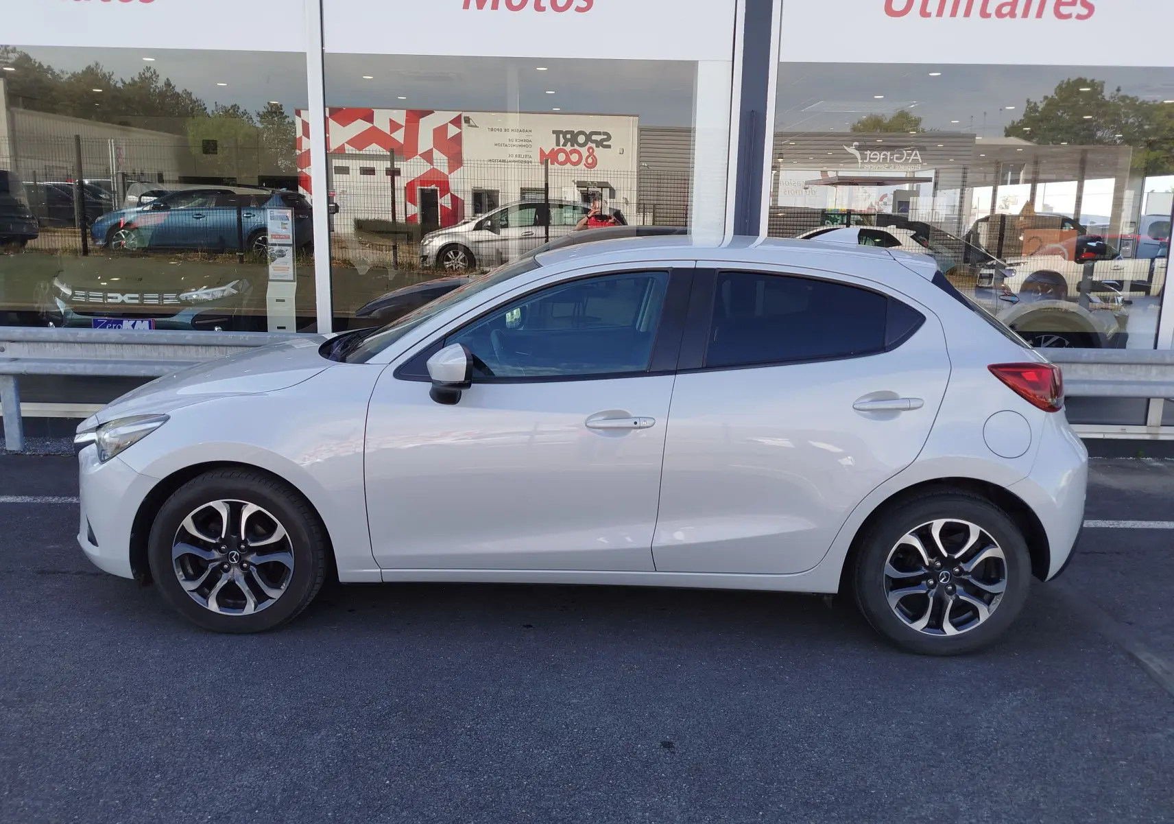 Vue de profil côté gauche d'une Mazda2 blanche nacrée avec jantes alliage bi-ton, stationnée devant une vitrine.