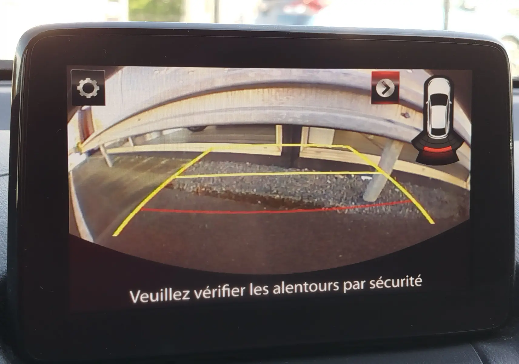 Écran de caméra de recul d'une Mazda2 blanche, montrant une barrière métallique vue de l'arrière du véhicule.