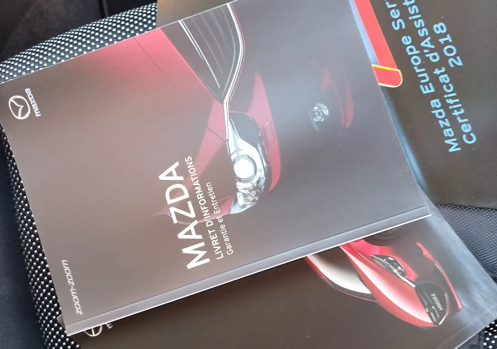 Image montrant des brochures Mazda2 posées sur un siège de voiture, avec une couverture présentant un avant de Mazda rouge en gros plan.