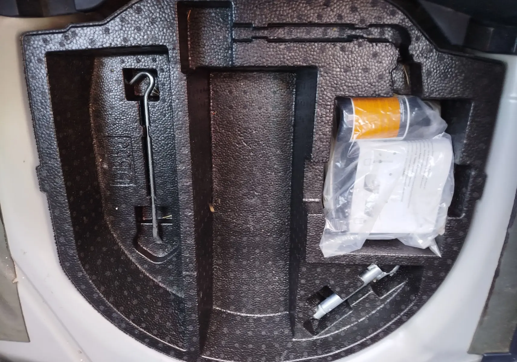 Vue plongeante sur le rangement en mousse noire dans le coffre d'une Mazda2 blanche, contenant des outils et un kit de réparation.