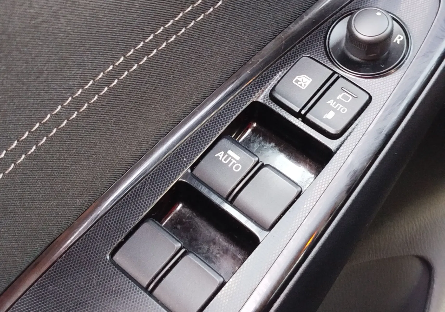 Gros plan sur les commandes de lève-vitres et réglage des rétroviseurs côté conducteur d'une Mazda2 blanche.