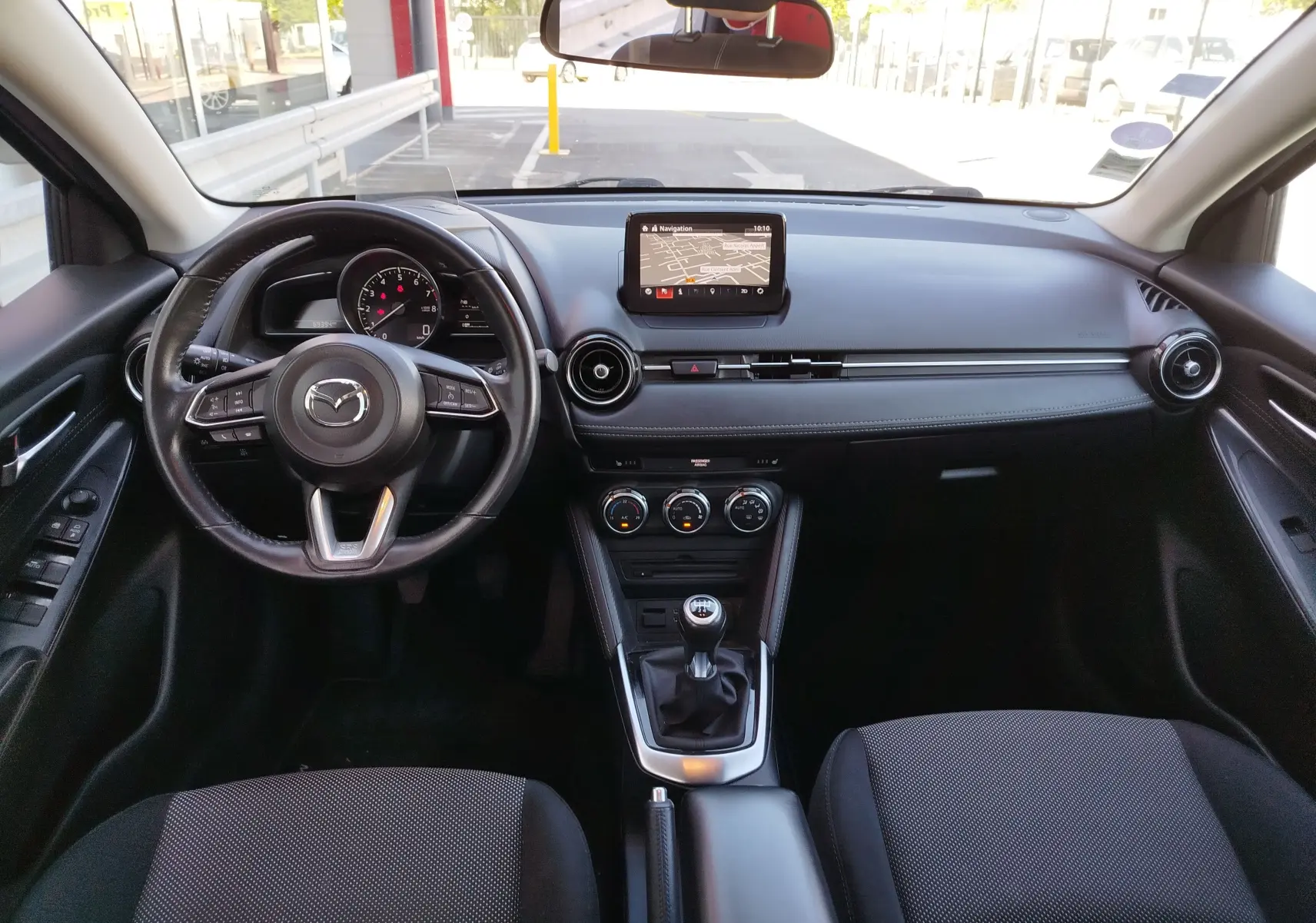 Vue intérieure avant de la Mazda2 2018 avec tableau de bord noir, écran central et boîte manuelle visible.