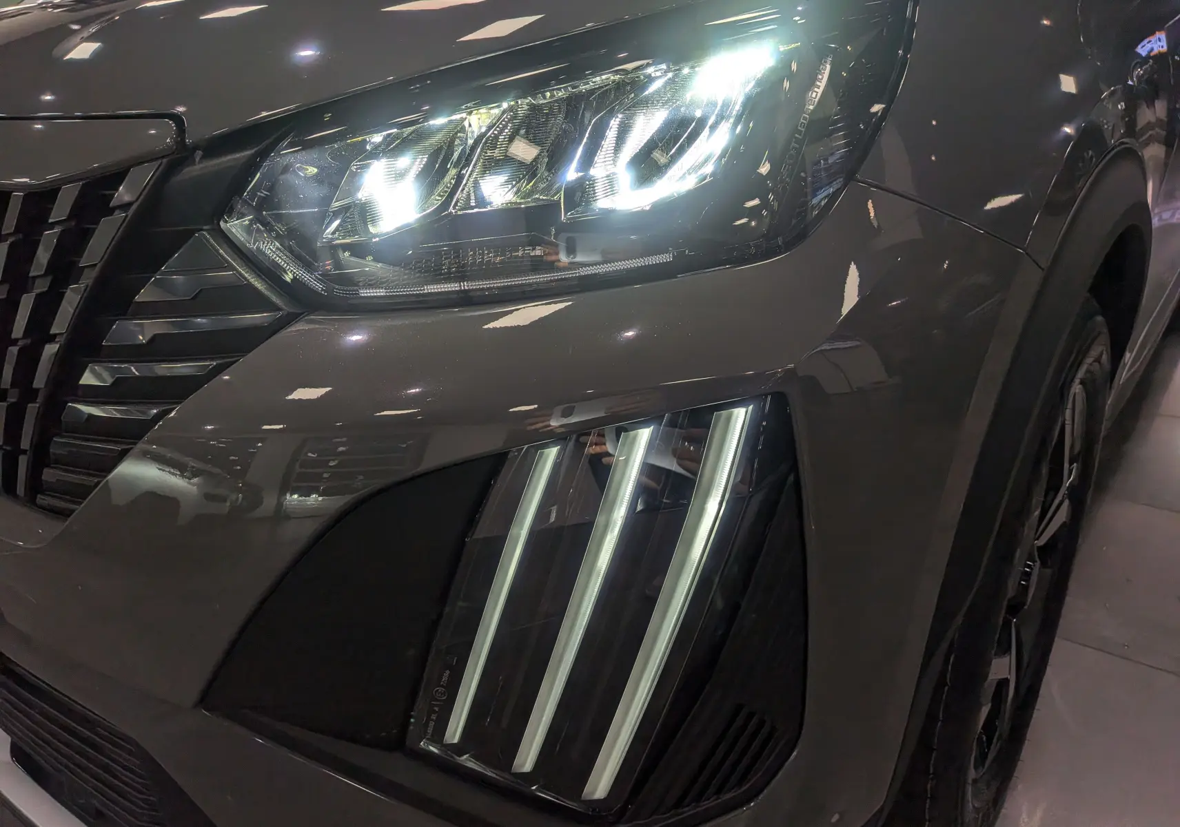 Gros plan sur le phare avant droit allumé du Peugeot 2008 Hybrid 145 gris Sélénium avec signature lumineuse verticale.