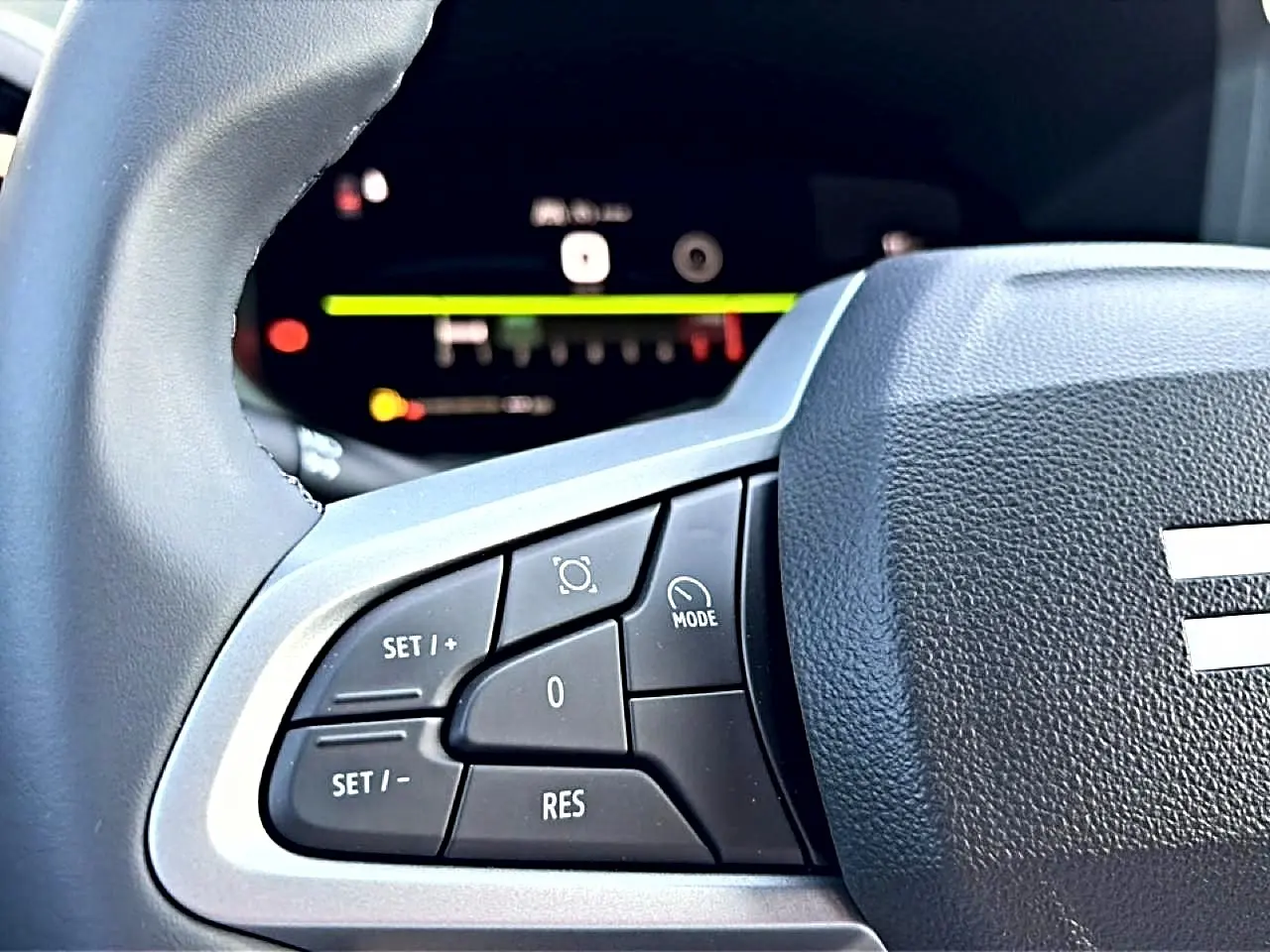 Gros plan sur les commandes multifonctions à gauche du volant noir du Dacia Bigster blanc, avec tableau de bord numérique flou en arrière-plan.
