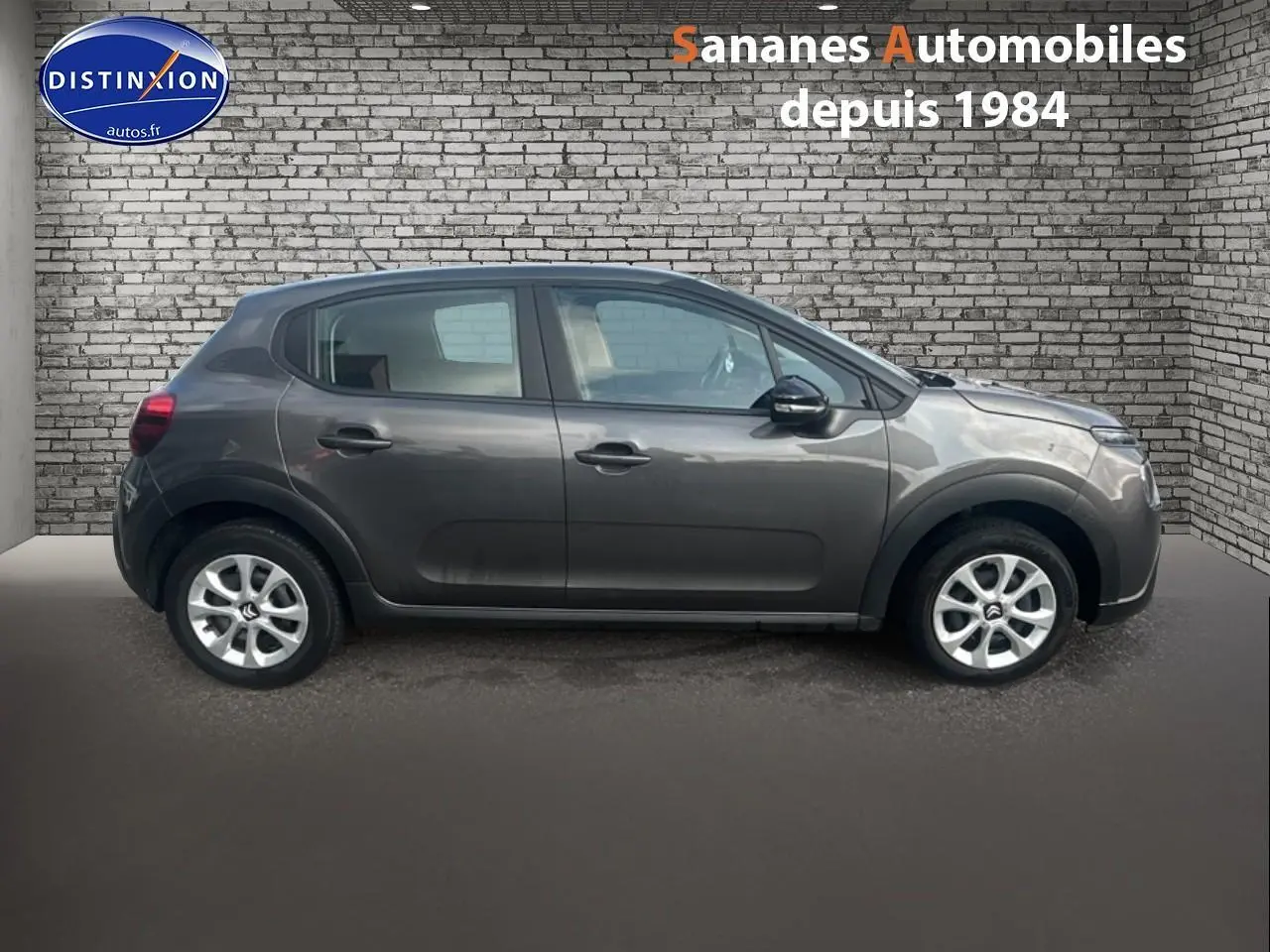 Profil côté gauche d'une Citroën C3 gris foncé 2023 avec jantes argentées, stationnée devant un mur en briques.