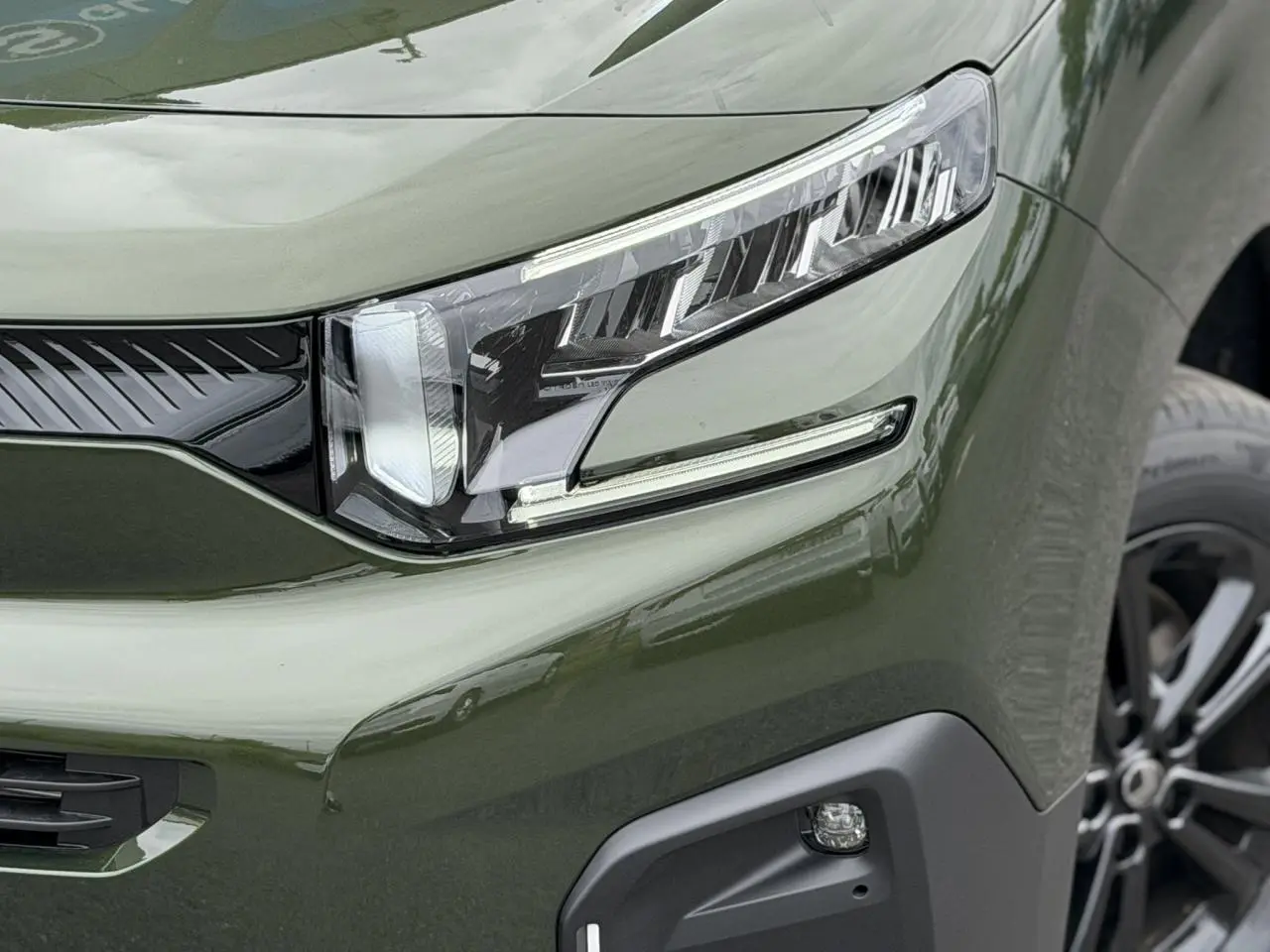Gros plan sur le phare avant droit et la calandre d’un Citroën Berlingo Multispace vert Sirkka avec jante alliage noire.
