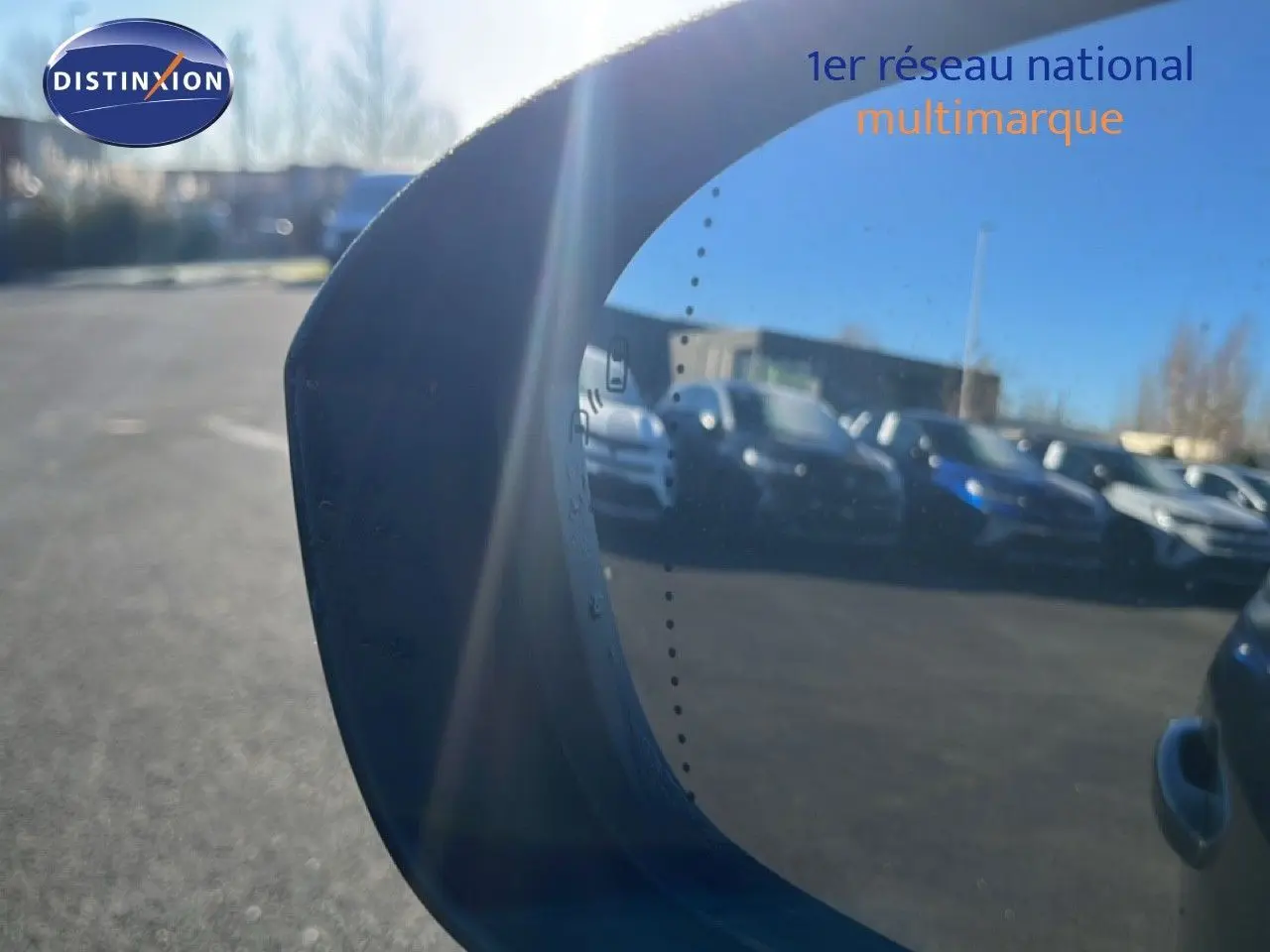 Rétroviseur gauche reflétant plusieurs voitures sur un parking par une journée ensoleillée.