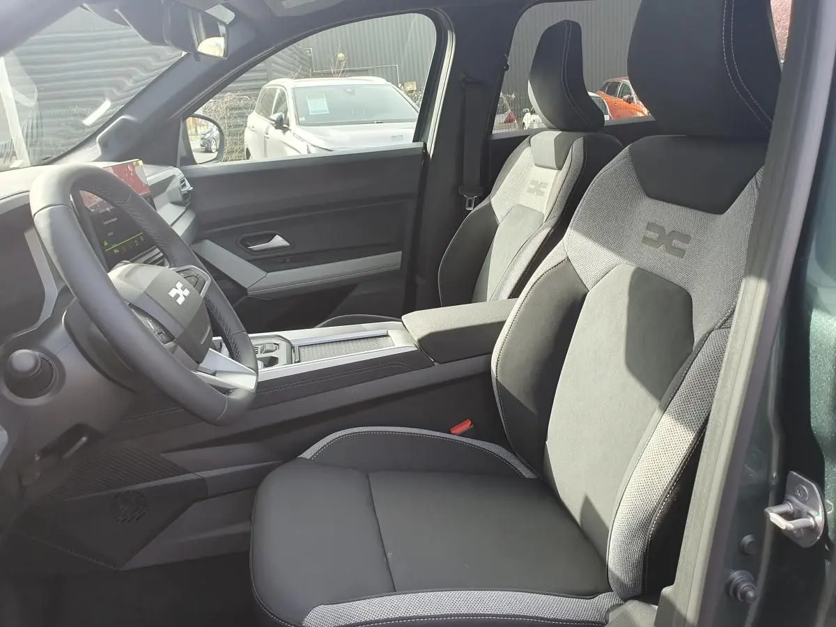 Vue intérieure côté conducteur du Dacia Bigster 2026 vert cèdre, sièges tissu gris avec logo brodé sur appuie-tête.
