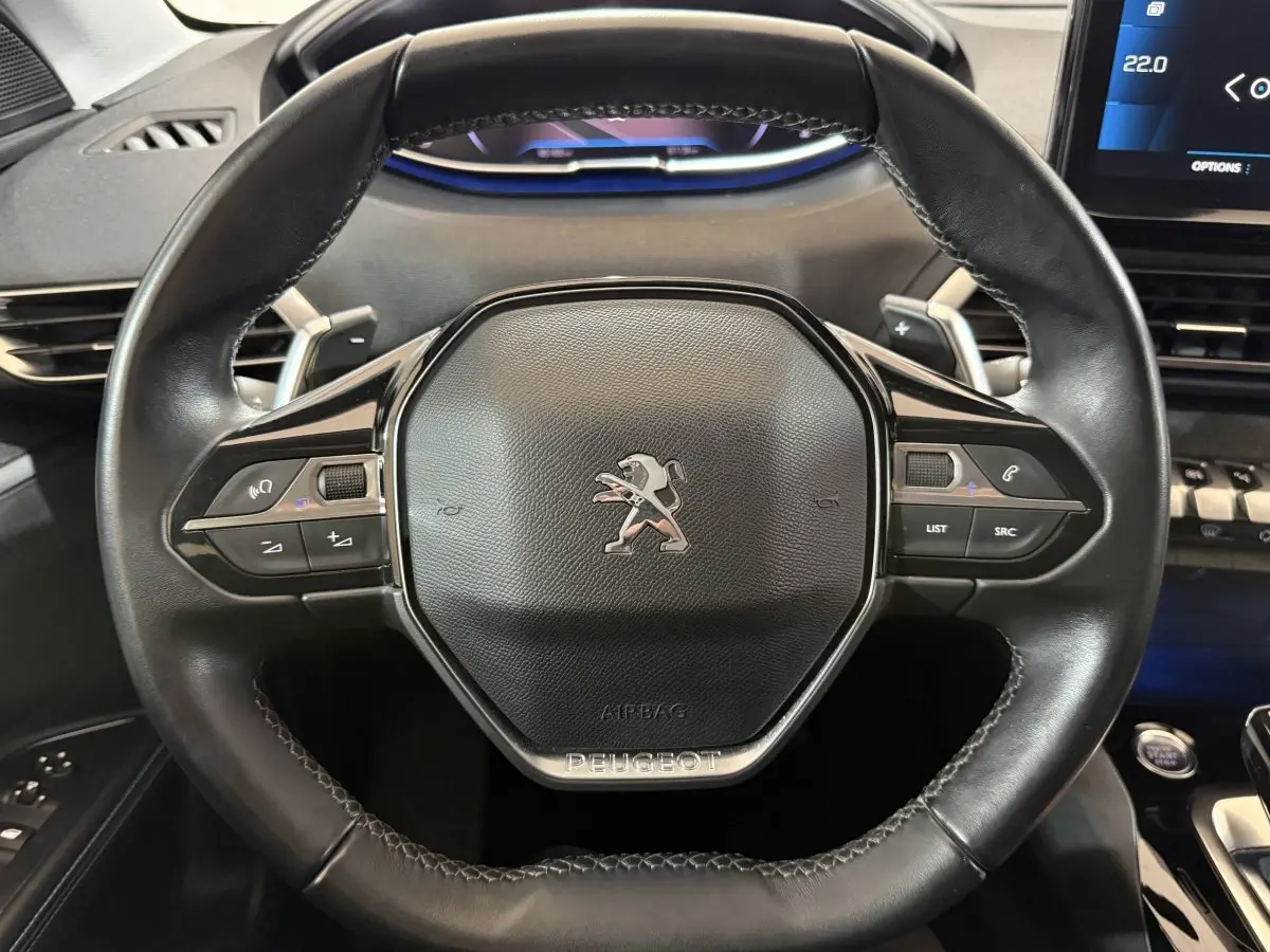 Gros plan sur le volant cuir noir du Peugeot 5008 2021 avec commandes intégrées et tableau de bord numérique bleu.