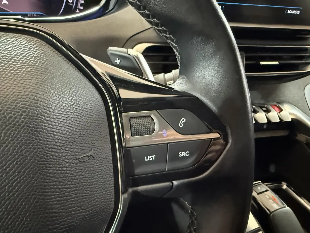 Gros plan sur le volant cuir noir du Peugeot 5008 2021, avec commandes audio et palette de changement de vitesse visible.