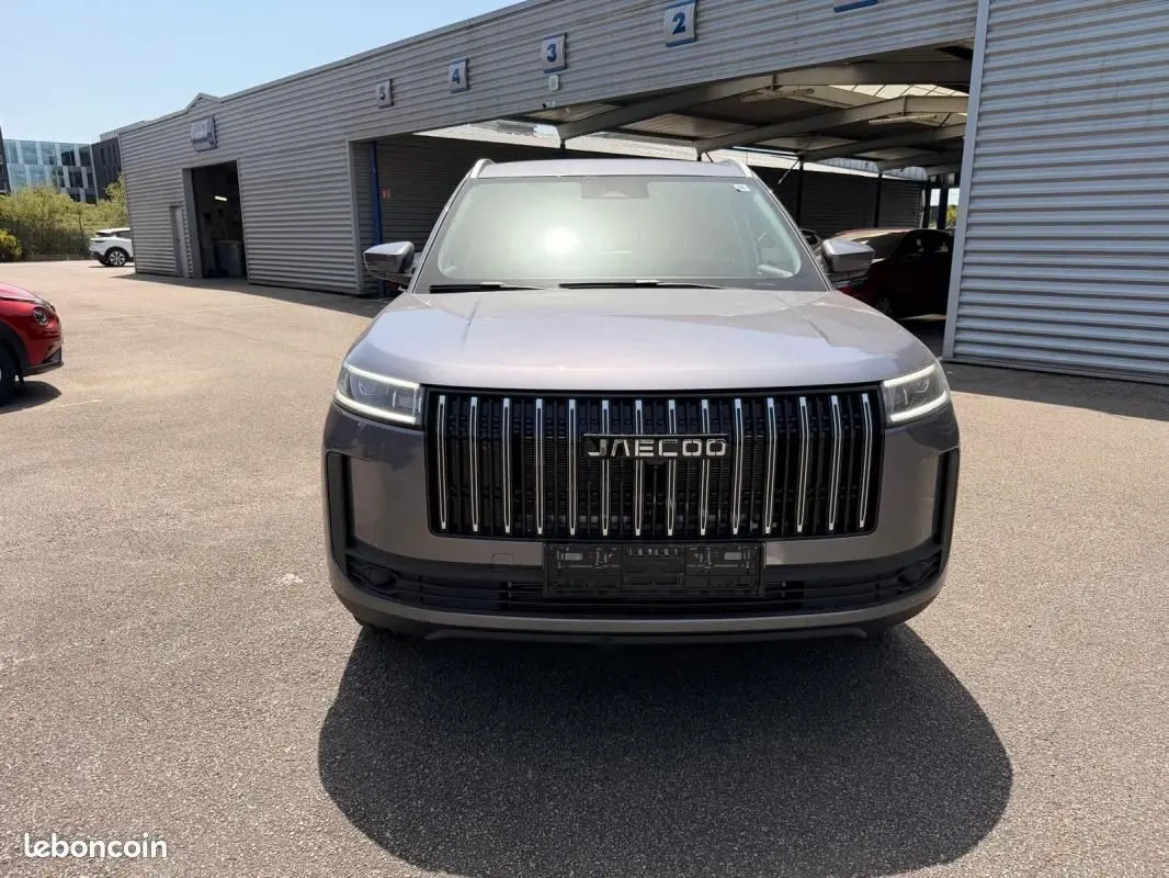 Vue 3/4 avant du JAECOO J5 gris Phantom avec calandre verticale noire et phares LED fins, stationné devant un bâtiment.