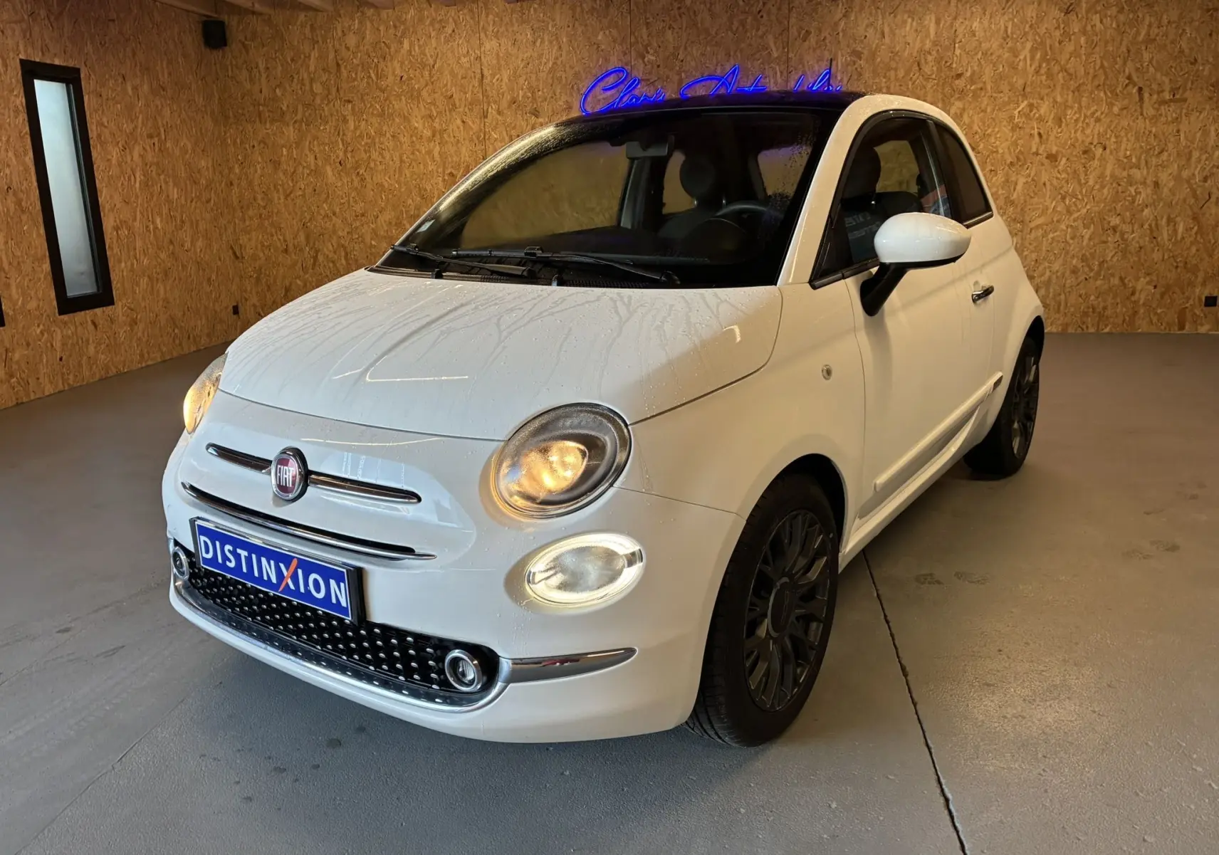 Vue 3/4 avant d'une Fiat 500 blanche de 2021 avec phares allumés et jantes foncées dans un garage.