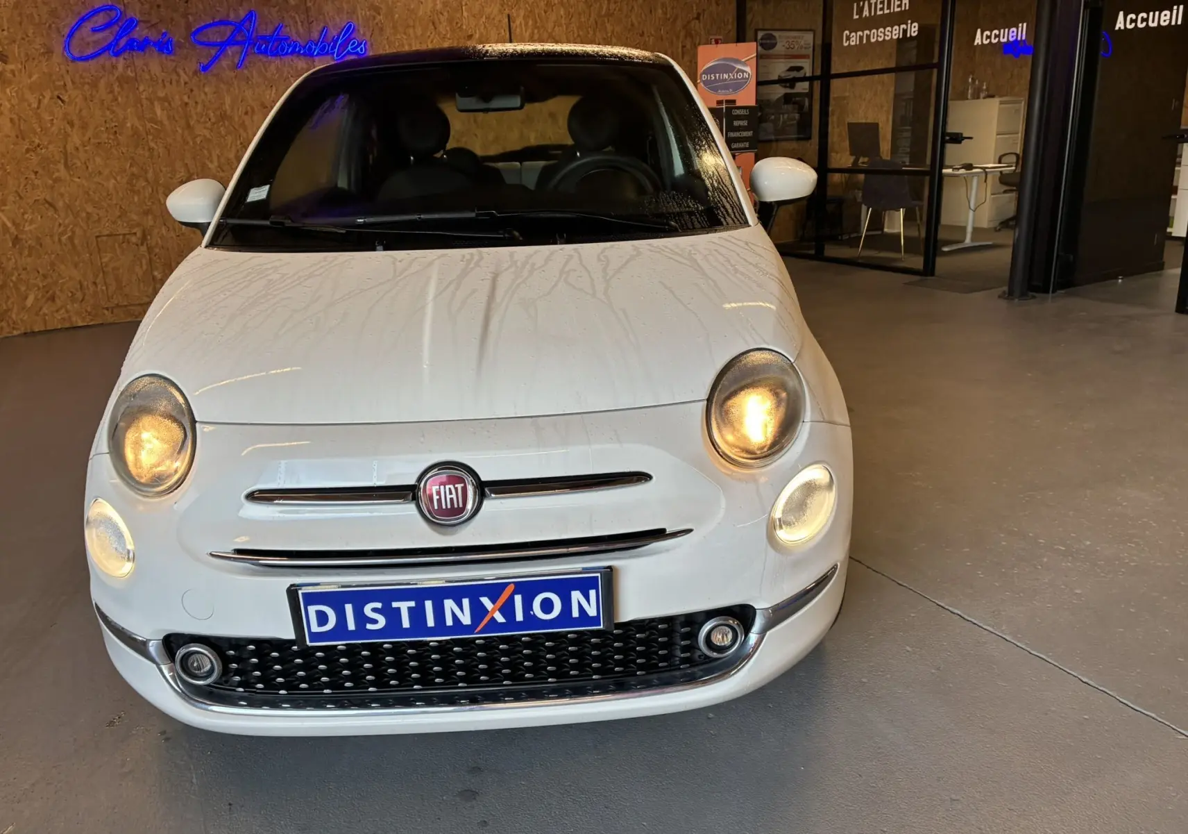 Vue avant d'une Fiat 500 blanche de 2021 avec phares allumés, stationnée dans un garage intérieur.