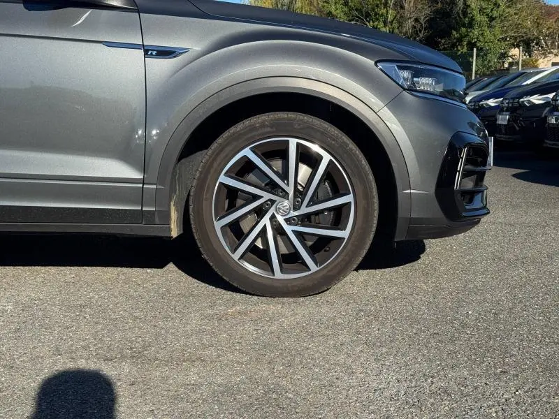 Gros plan sur la roue avant droite et partie avant grise métallisée du Volkswagen T-Roc 2.0 TSI R avec jante alu distinctive.