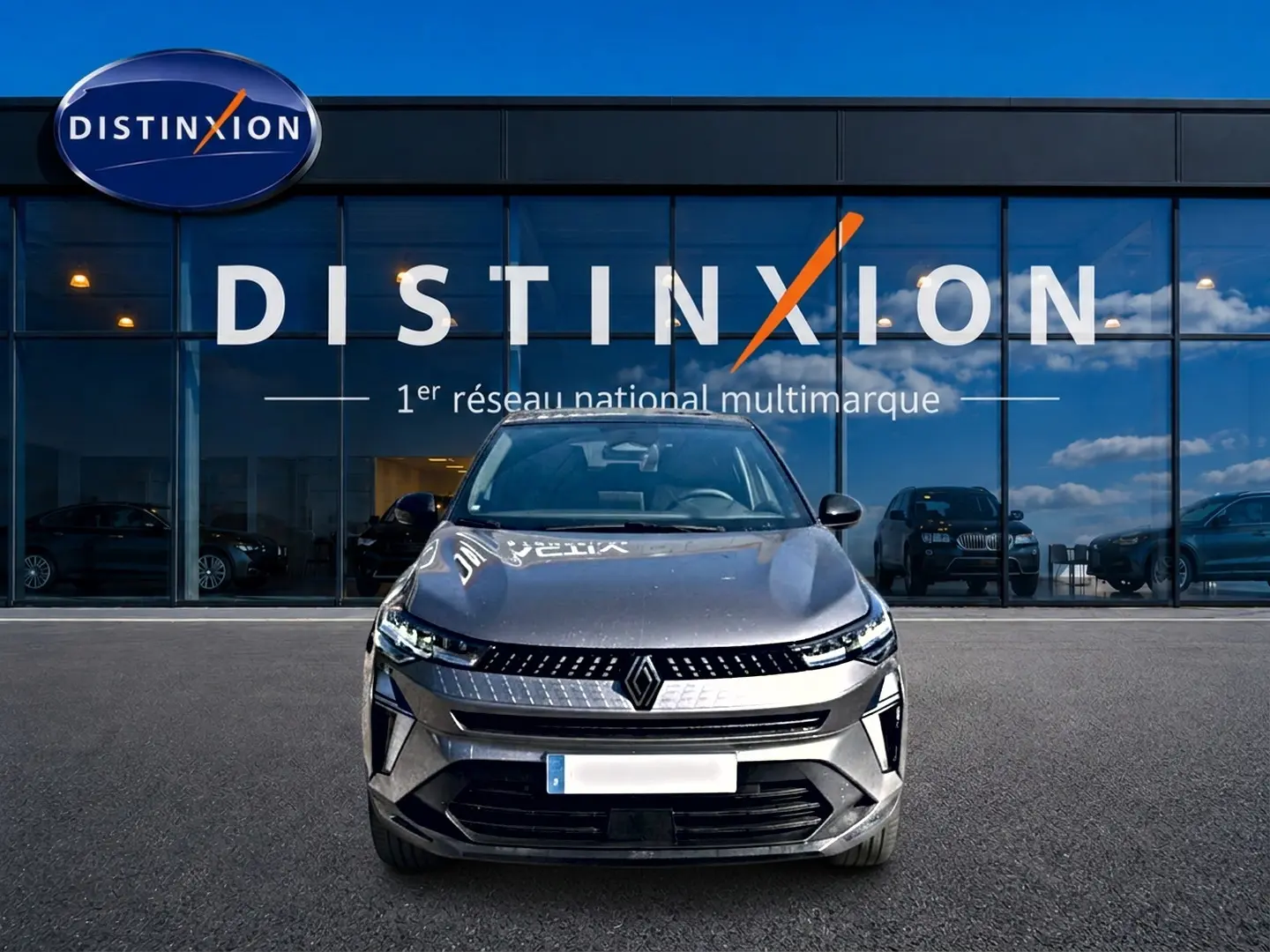 Vue frontale d'un Renault Captur gris cassiopée 2026, avec calandre moderne et phares LED allumés, devant une vitrine Distinxion.