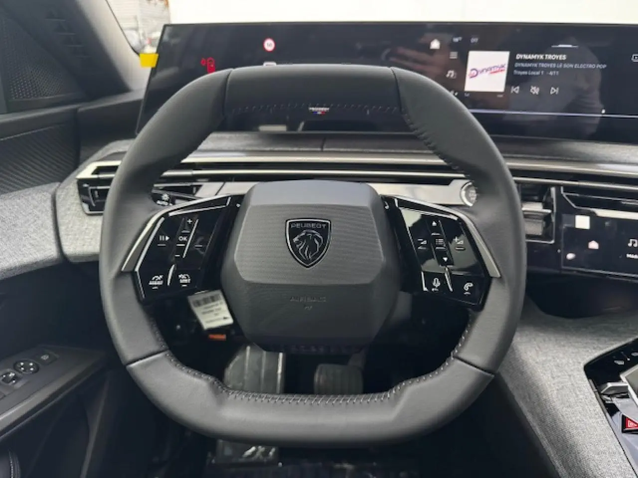 Volant compact cuir noir avec logo Peugeot, tableau de bord numérique panoramique et habillage tissu gris clair visible depuis le poste de conduite