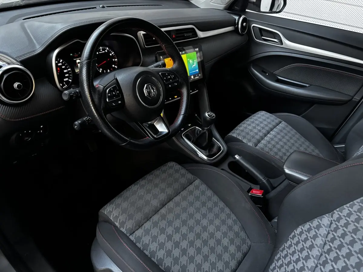 Vue intérieure côté conducteur du MG ZS 2022, volant cuir, sièges tissu gris à motifs, écran tactile central allumé.