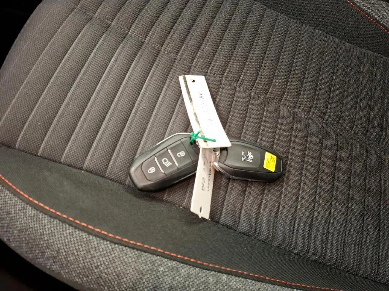 Clé de voiture Peugeot posée sur siège tissu noir avec surpiqûres orange, vue rapprochée intérieure.