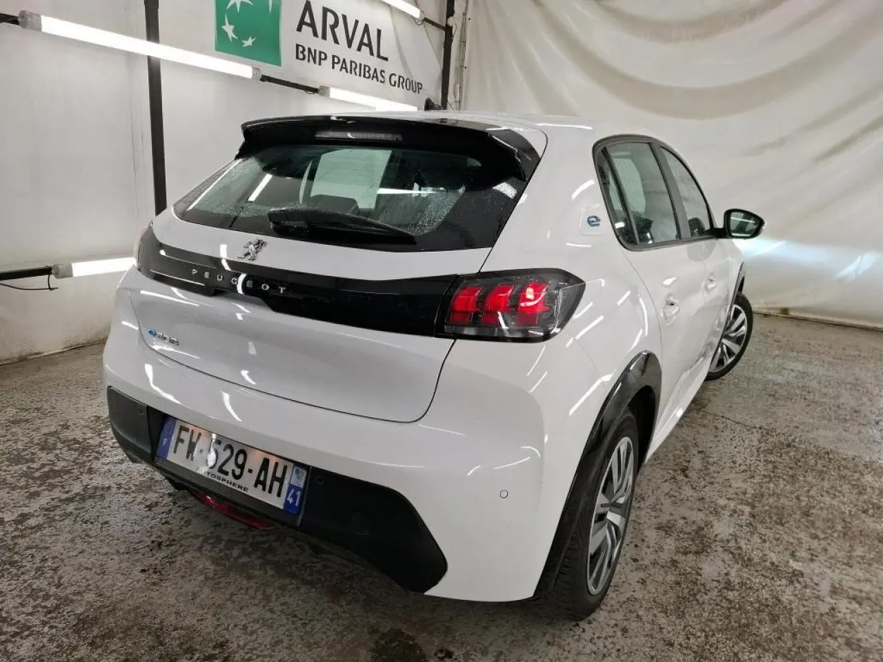 Vue 3/4 arrière droite d'une Peugeot 208 électrique blanche avec bandeau noir brillant entre feux arrière dans un garage.