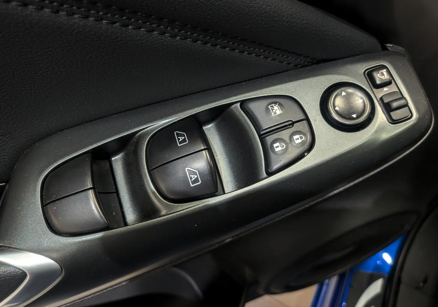 Gros plan sur les commandes de lève-vitres et rétroviseurs côté conducteur du Nissan Juke Hybrid bleu magnétique avec intérieur noir.