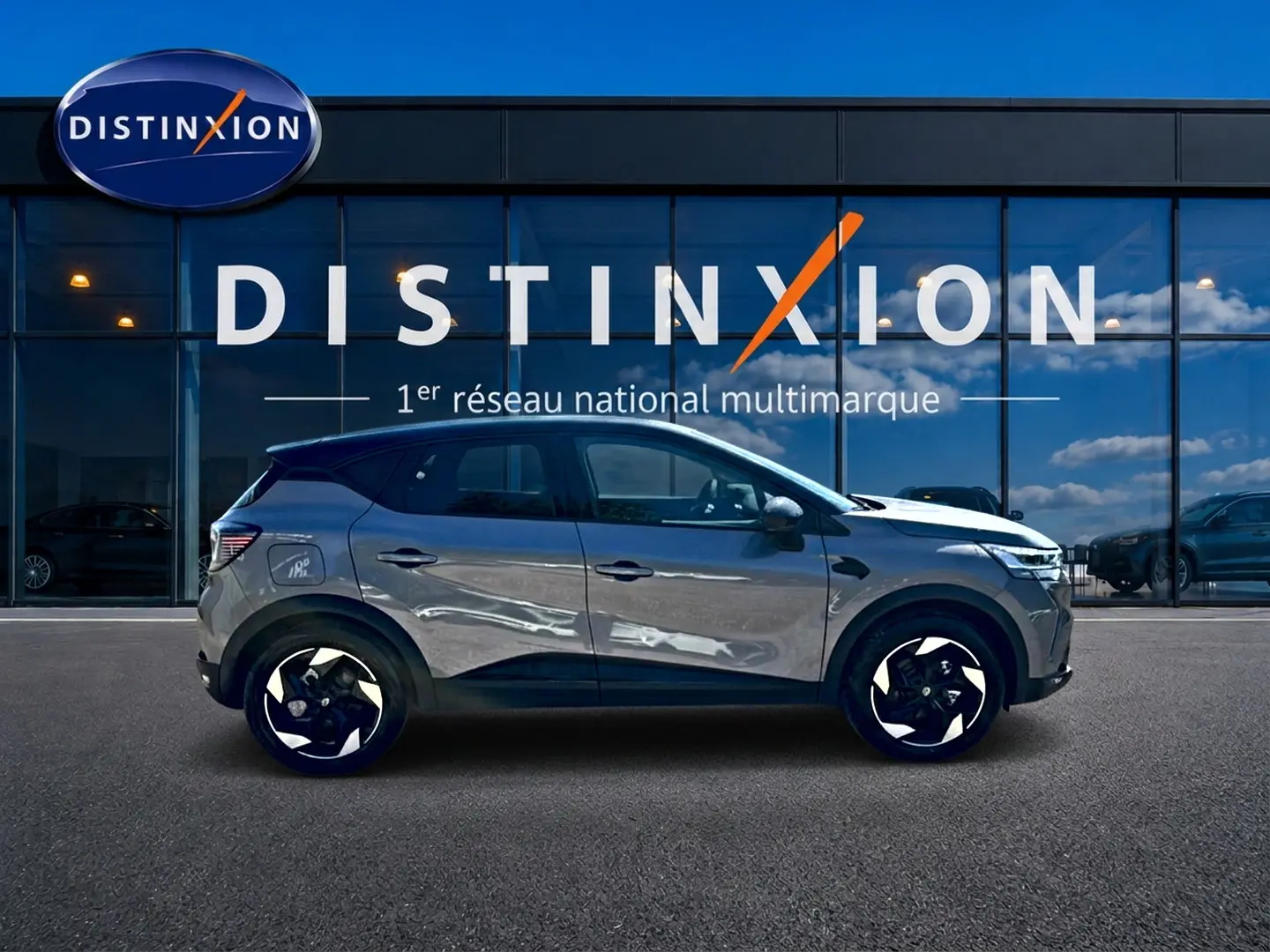Profil droit d’un Renault Captur gris cassiopée 2026 devant une vitrine Distinxion, avec jantes alu noires et blanches.