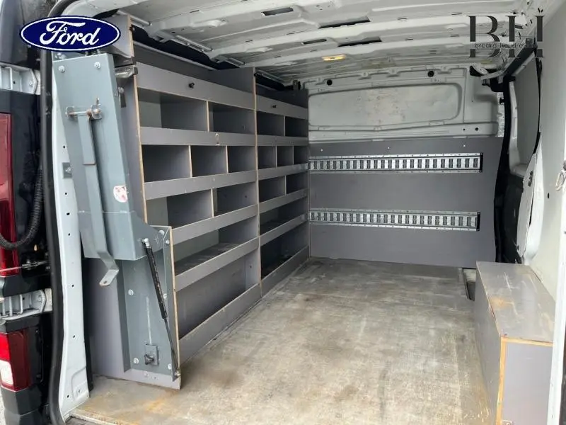 Vue intérieure arrière du fourgon Renault Trafic blanc glacier, montrant les étagères de rangement grises et le plancher usé.