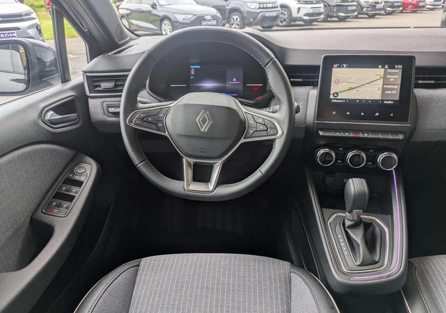 Vue intérieure avant centrée sur le volant et tableau de bord de la Renault Clio hybride 2025 en noir étoile métal, avec écran tactile et levier de vitesses.