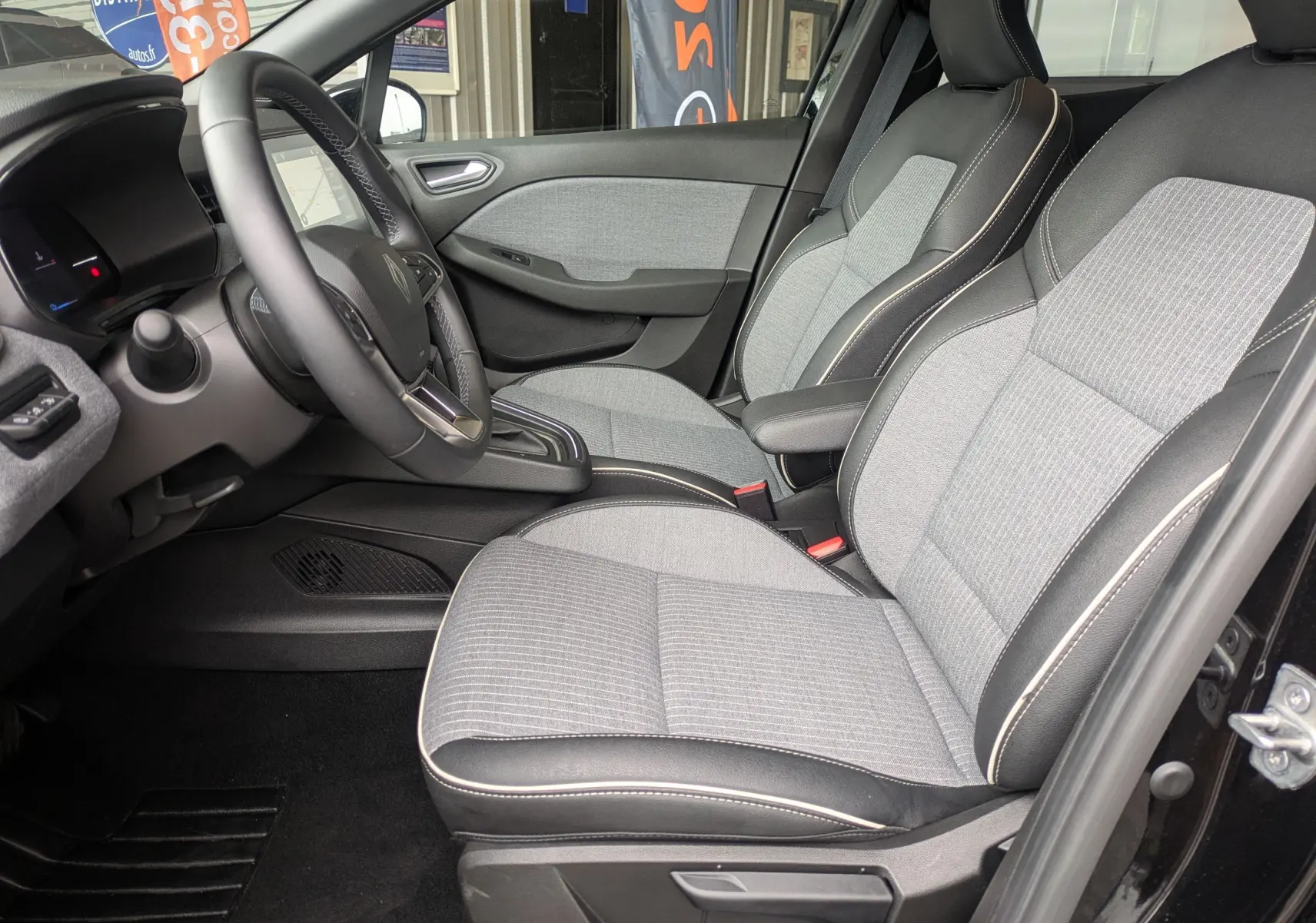 Vue intérieure côté gauche de la Renault Clio 2025, sièges tissu gris clair avec surpiqûres blanches et volant cuir noir.
