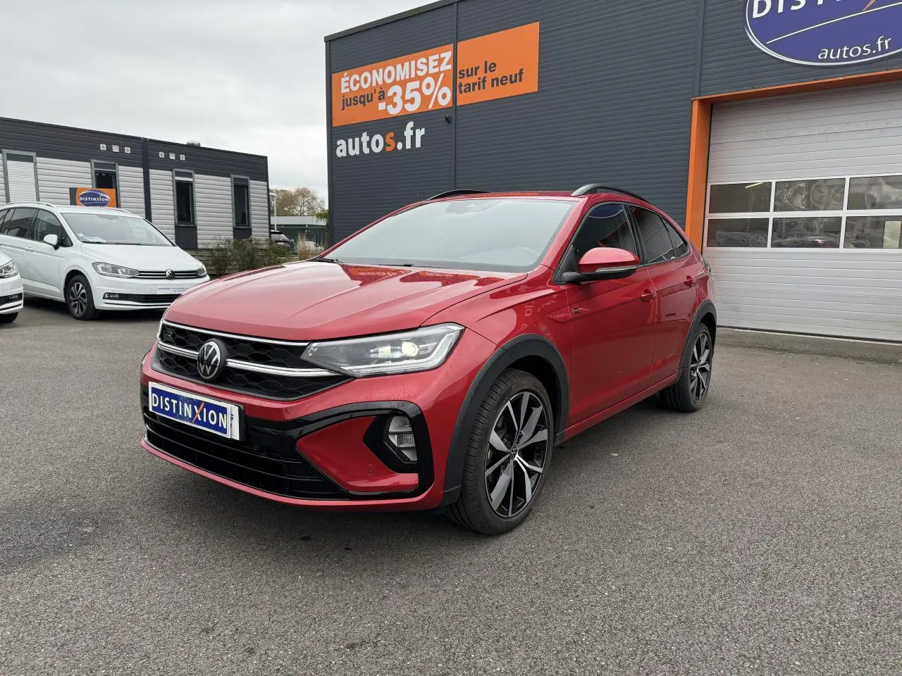 Volkswagen Taigo Rouge Roi en 3/4 avant droit, jantes noires brillantes, feux LED allumés, devant un garage autos.fr.