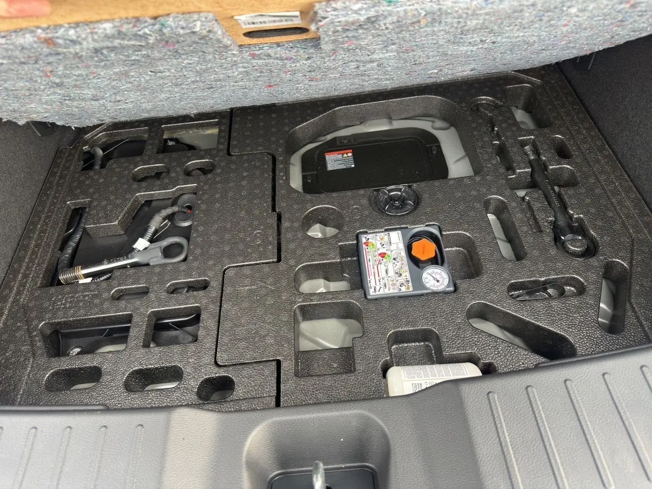 Coffre ouvert du Nissan Juke 2025 gris squale, vue plongeante sur le rangement d’outils et compresseur intégré.