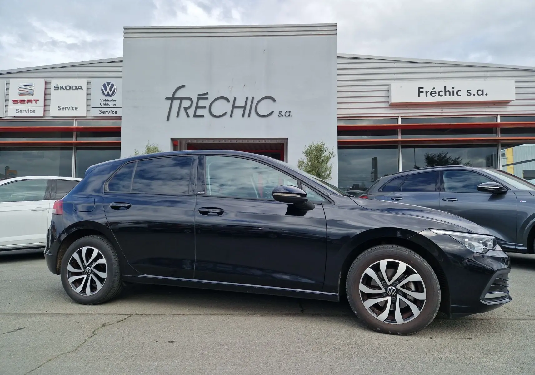 Profil côté gauche de la Volkswagen Golf 8 noire intense métal, avec jantes alliage et vitres teintées, devant un bâtiment commercial.