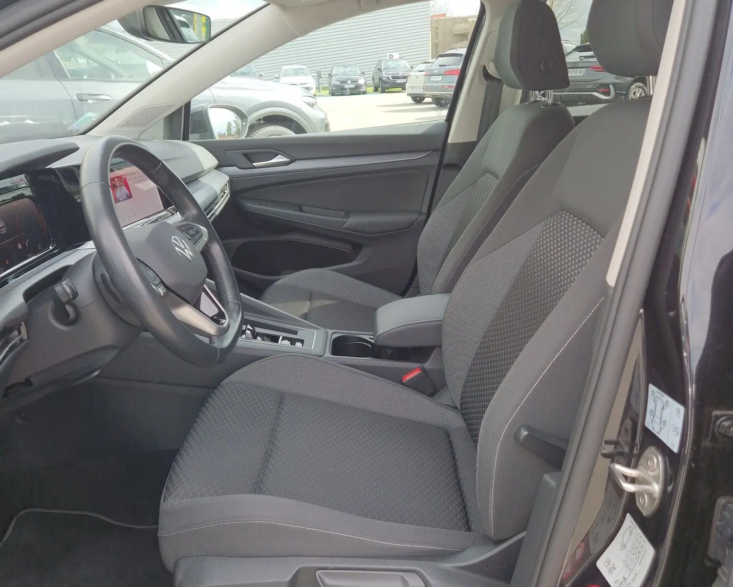 Vue intérieure côté conducteur de la Volkswagen Golf 8 noire, montrant les sièges avant et le tableau de bord moderne.