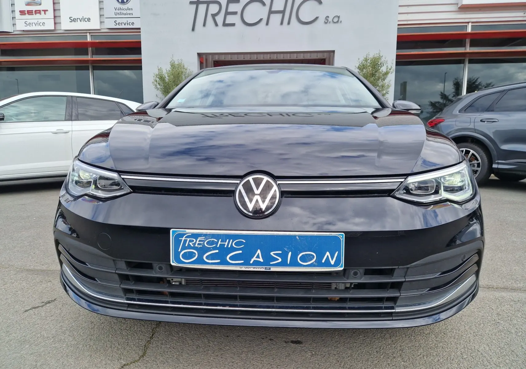 Vue frontale d'une Volkswagen Golf 8 noire intense métal, avec calandre et phares LED distinctifs, devant un concessionnaire.