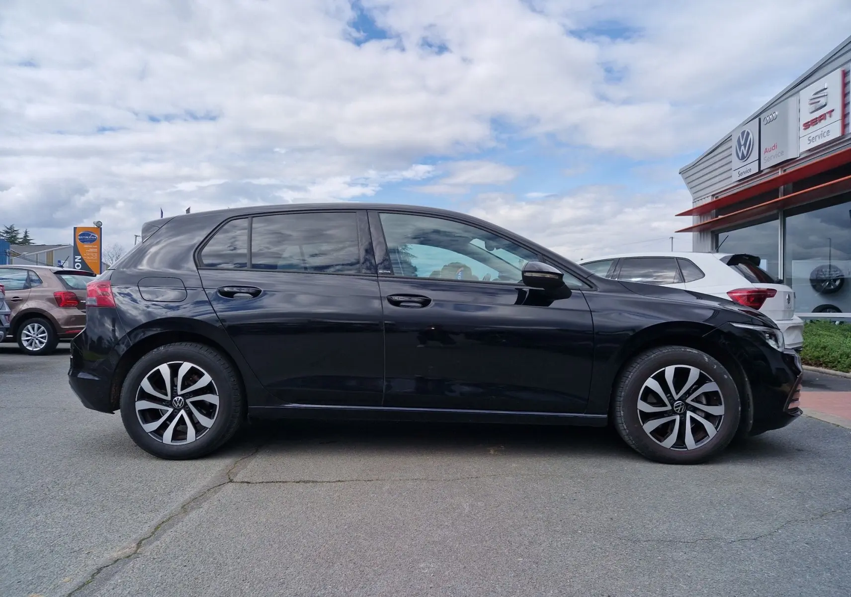 Vue latérale droite d'une Volkswagen Golf 8 noire intense métal, avec jantes alliage distinctives, stationnée devant un concessionnaire.
