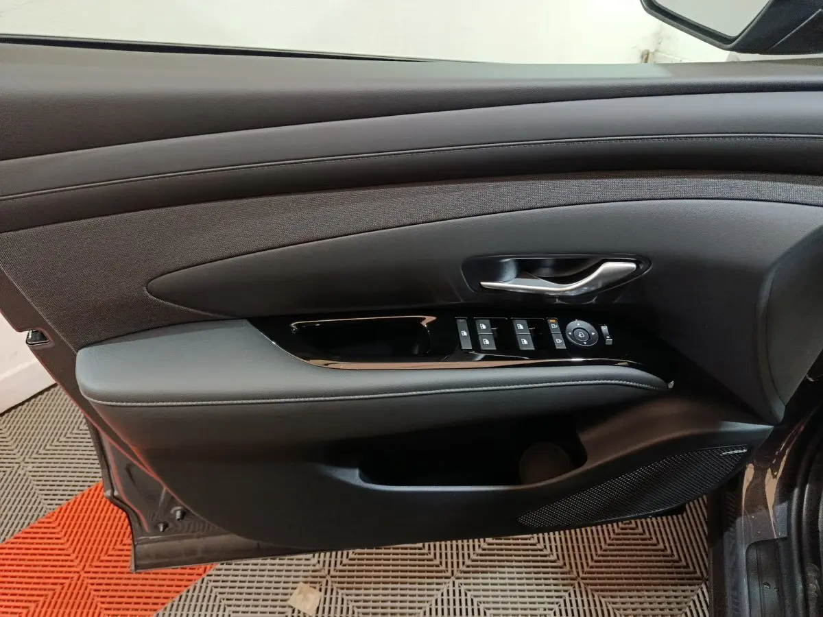Vue intérieure du panneau de porte côté conducteur du Hyundai Tucson gris foncé, avec commandes électriques des vitres et poignée argentée.