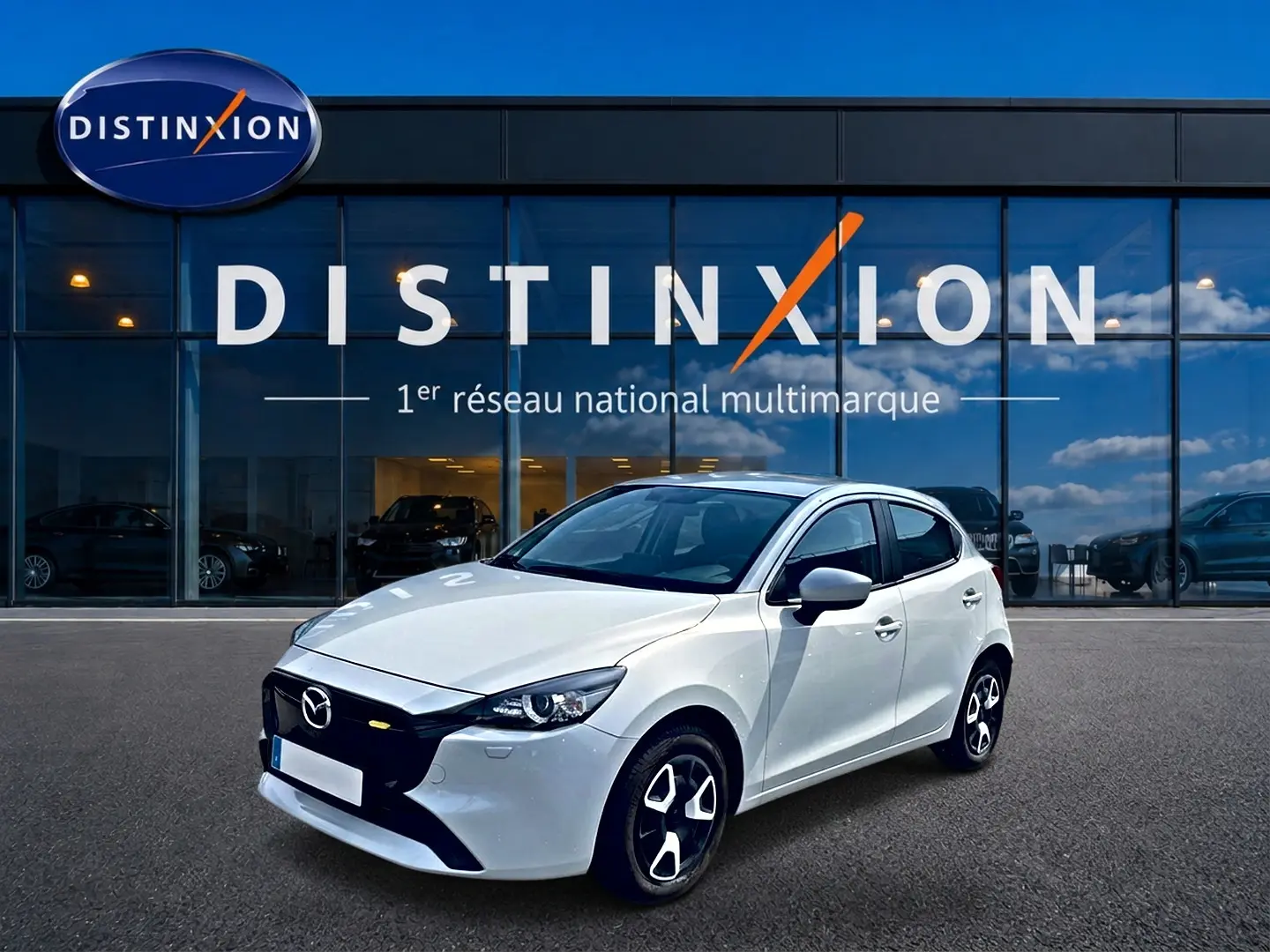 Vue 3/4 avant d'une Mazda2 blanche 2024 avec calandre noire et jantes distinctives, devant une vitrine Distinxion.