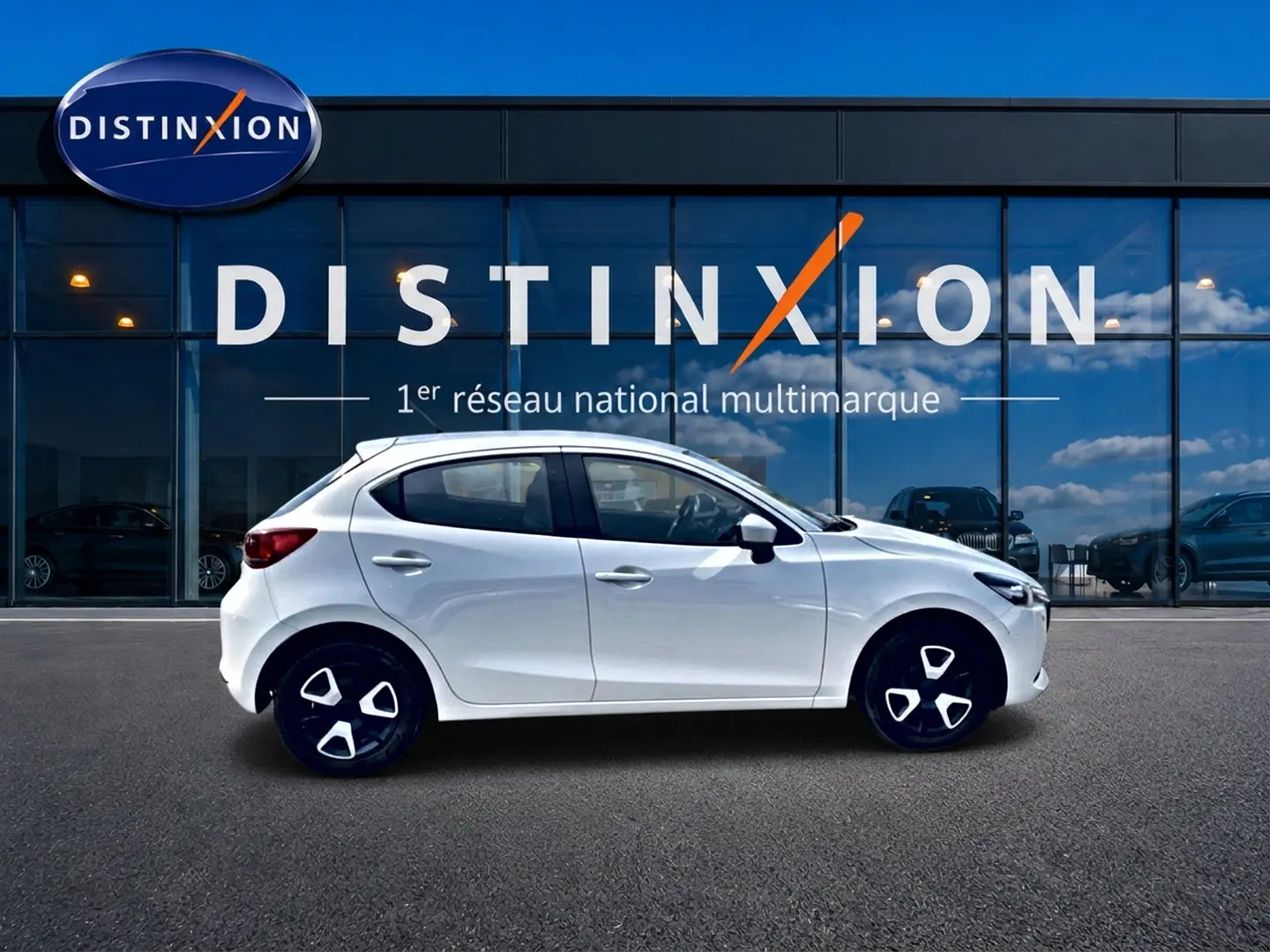 Vue latérale droite d'une Mazda2 blanche 2024 avec jantes noires et détails argentés, devant une vitrine Distinxion.