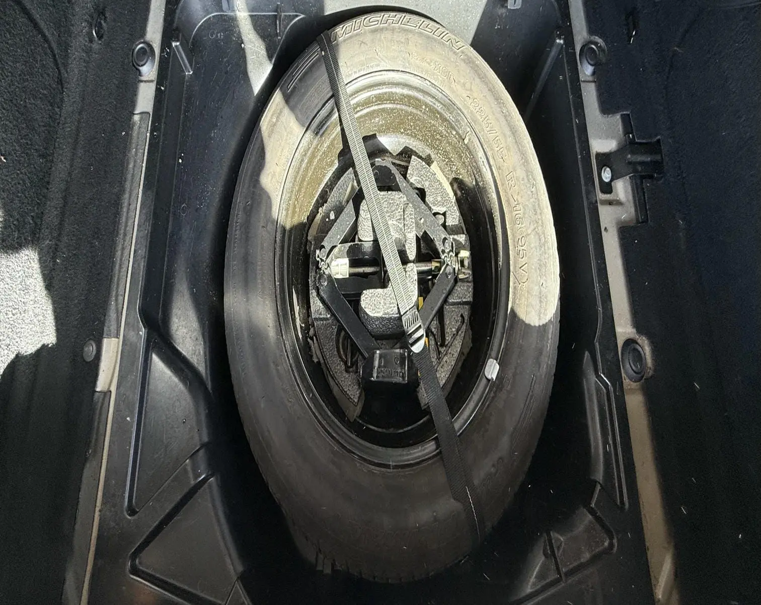 Vue plongeante sur la roue de secours Michelin et son équipement de levage dans le coffre d'une Peugeot 607 grise.