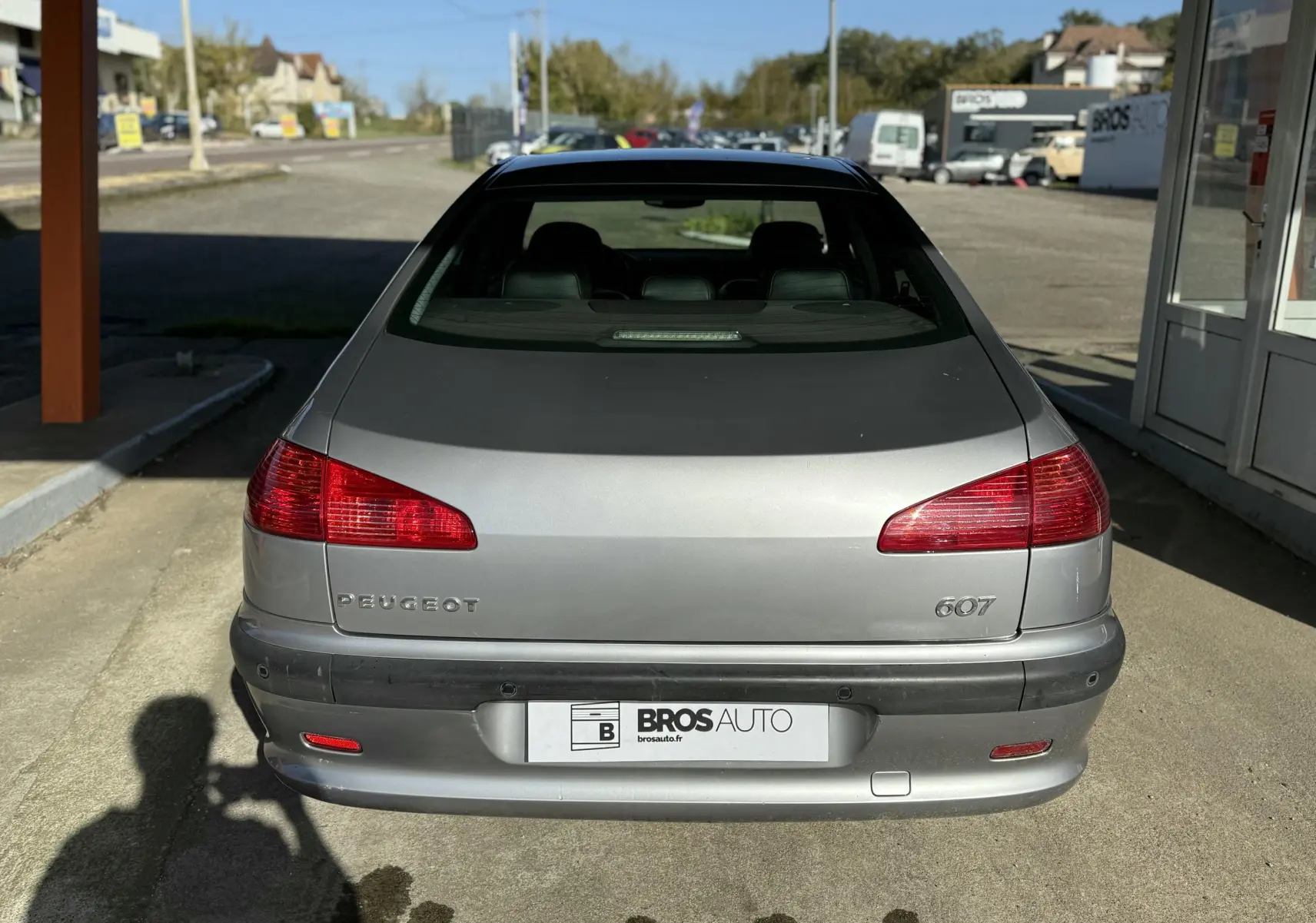Vue arrière d'une Peugeot 607 gris métallisé garée devant un bâtiment commercial par temps ensoleillé.