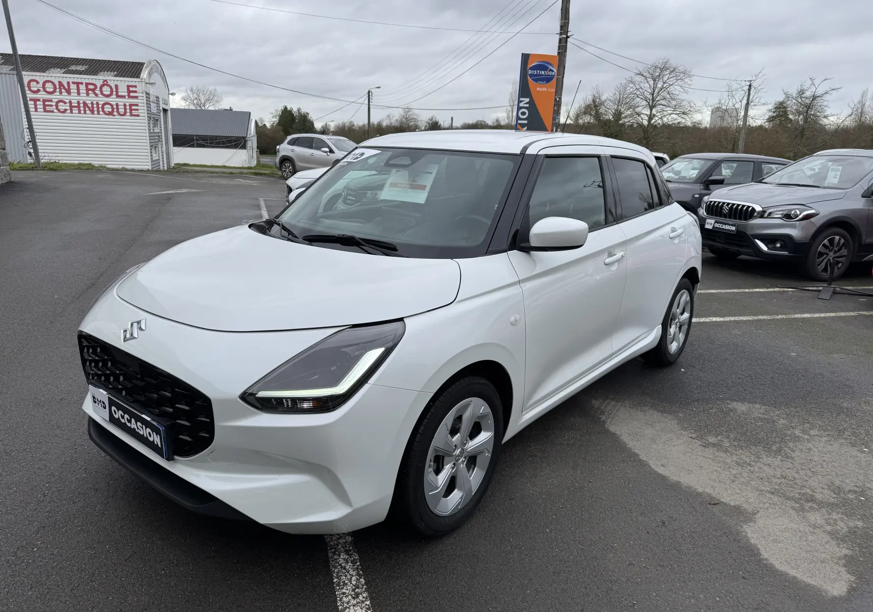 Vue 3/4 avant droite d'une Suzuki Swift 1.2 Privilège Hybrid blanche, avec calandre noire et jantes alliage.